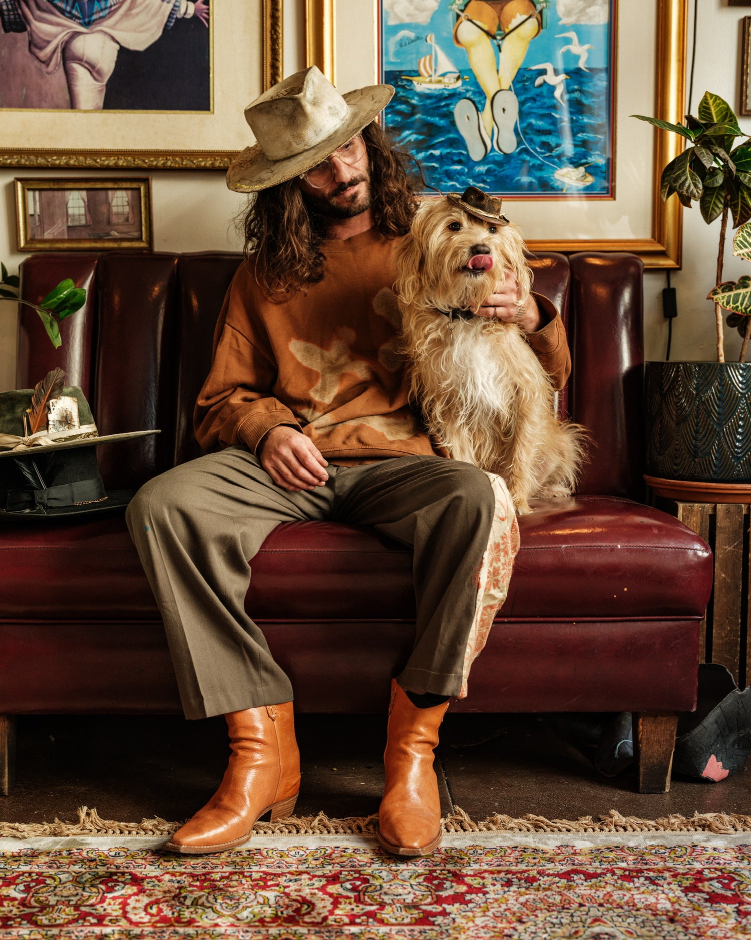 A man with long curly hair wearing a white wide brim hat sitting on a red restaurant booth with a dog and wall art behind him.