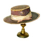 An antique Victorian-style hat with a dark brown brim, a patterned gold and purple crown, and a cream ribbon band, displayed on a gold-colored metal stand against a white background.
