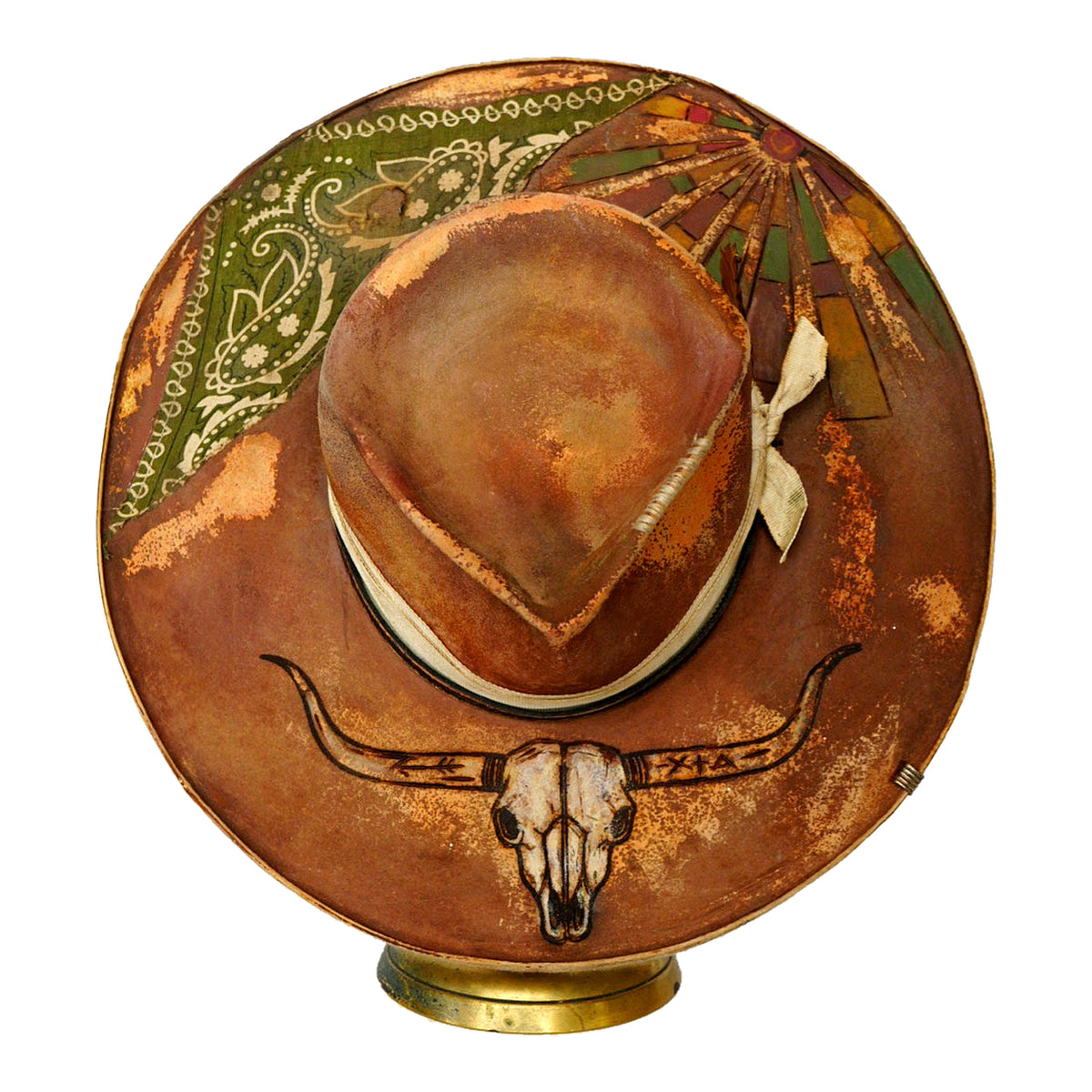 A brown cowboy hat with hand-painted rustic western designs, including a skull motif on the brim and a green paisley pattern on the crown, displayed on a small gold-colored stand against a white background.